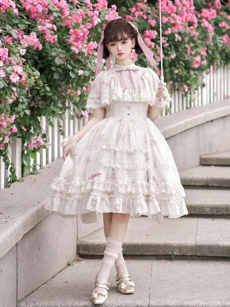 a young woman in a flowing white and pink floral lolita dress poses in a garden setting. she is standing on stone steps with a white picket fence and pink roses behind her. the lighting is soft and natural, highlighting the delicate details of her dress.