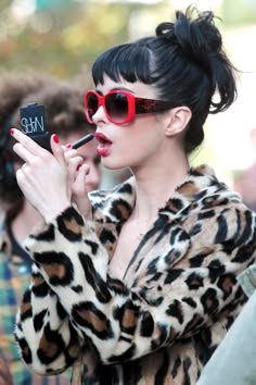 a young woman with black hair styled in an updo, is wearing large red sunglasses, a leopard print coat, and is applying red lipstick. she looks away from the camera, towards her left, and is smiling.