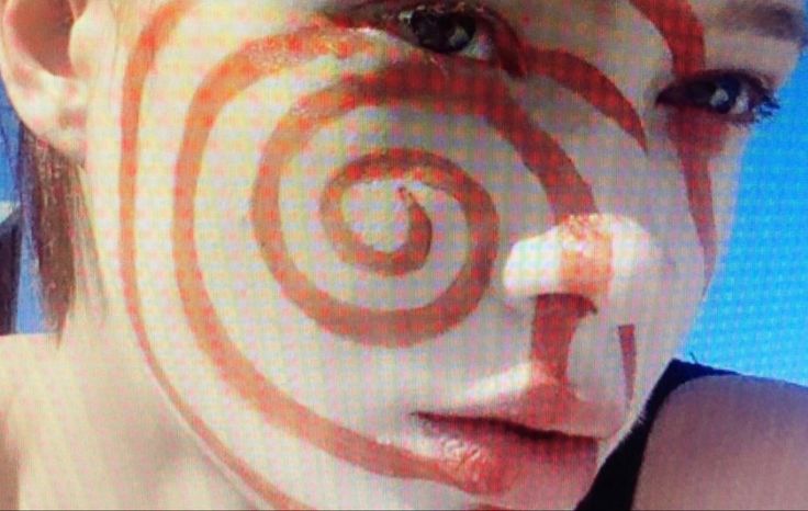 this is a closeup image of a woman's face. she is wearing red makeup on her face in a spiral pattern, with blue eyeshadow on her eyes. the background is blue. the image is focused on her face, and she is looking at the camera.