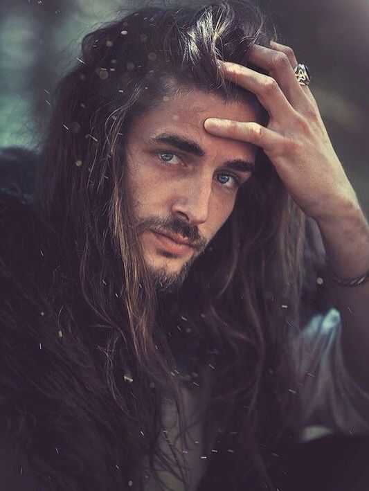 a portrait of a man with long brown hair, a beard, and blue eyes. he is touching his forehead with his right hand, which has a ring on it. he is wearing a dark garment. the background is blurry and out of focus.