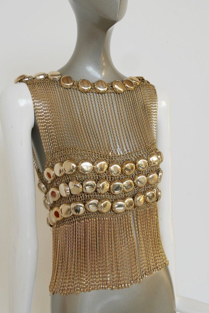 a gold chainmail top is displayed on a mannequin. the top features a series of metallic beads around the neckline and three rows across the upper chest. gold chainmail fringes hang from the lowest row of beads, creating a layered effect.