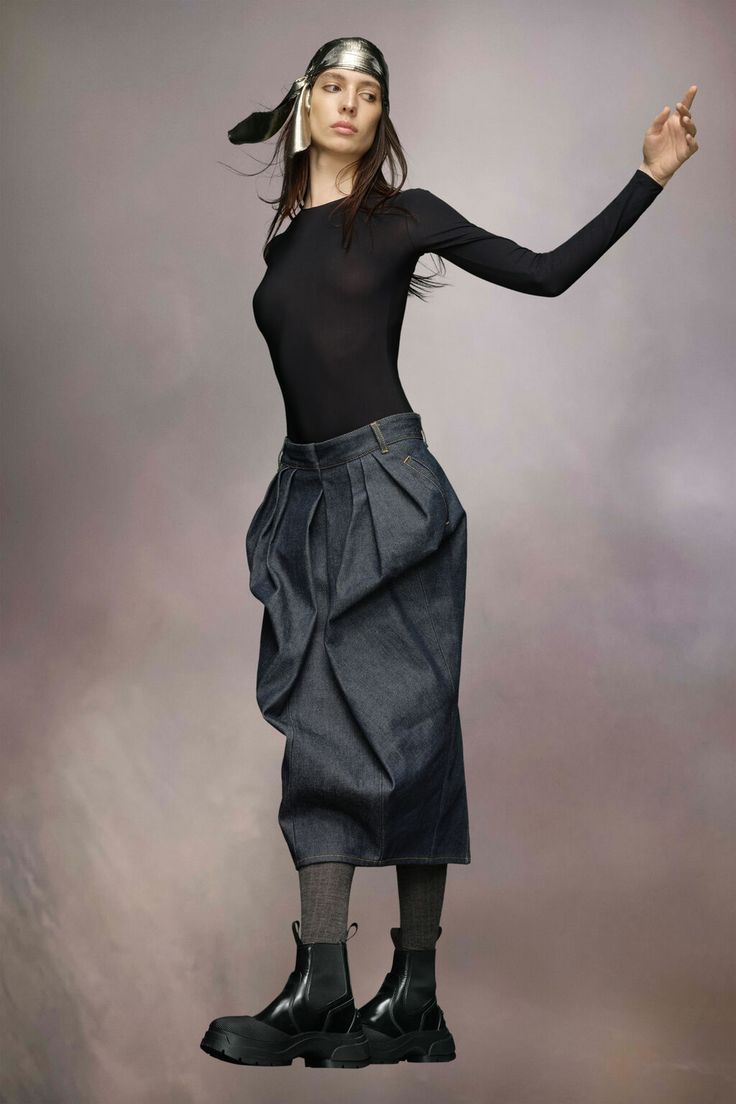 a fulllength shot of a fashion model wearing a black longsleeve bodysuit, a pleated denim midi skirt, black ankle boots, and a silver headband. the model is posing against a soft grey background, with her arm raised. the overall style is contemporary and minimalist.