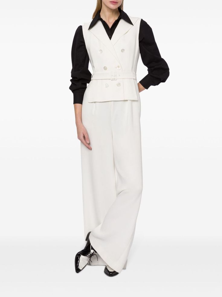 a woman in a white pantsuit, wearing a doublebreasted vest and a black blouse, is standing with her legs crossed. she is wearing black and white shoes.