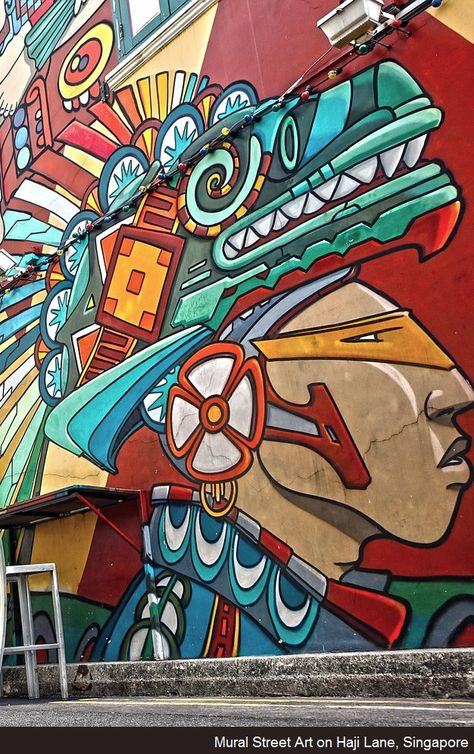 the image showcases a vibrant and intricately detailed mural on a building in haji lane, singapore. the artwork depicts a stylized head and headdress reminiscent of aztec or mayan indigenous art.  bold colors—teal, orange, red, yellow, blue, and green—dominate the piece, creating a strong visual impact.  the headdress features a complex design with geometric patterns and elements suggesting a mythical creature or mask. the style is a blend of traditional indigenous aesthetics and contemporary graphic design. the mural's texture appears somewhat rough, perhaps reflecting the building's surface. a portion of a street and a metal chair are visible in the bottom left corner, grounding the artwork within its urban setting. the overall feeling is energetic and culturally rich, reflective of haji lane's known artistic character.