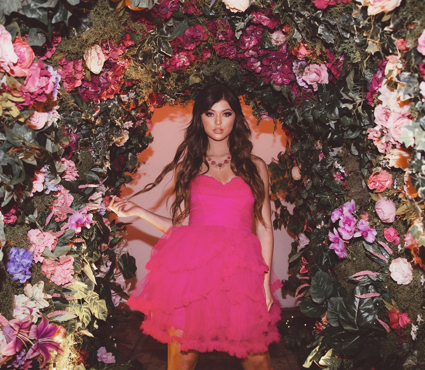 a woman wearing a pink tulle dress and necklace is posing in front of a floral archway. the archway is adorned with a variety of colorful flowers and greenery. the woman has long brown hair and is looking at the camera.