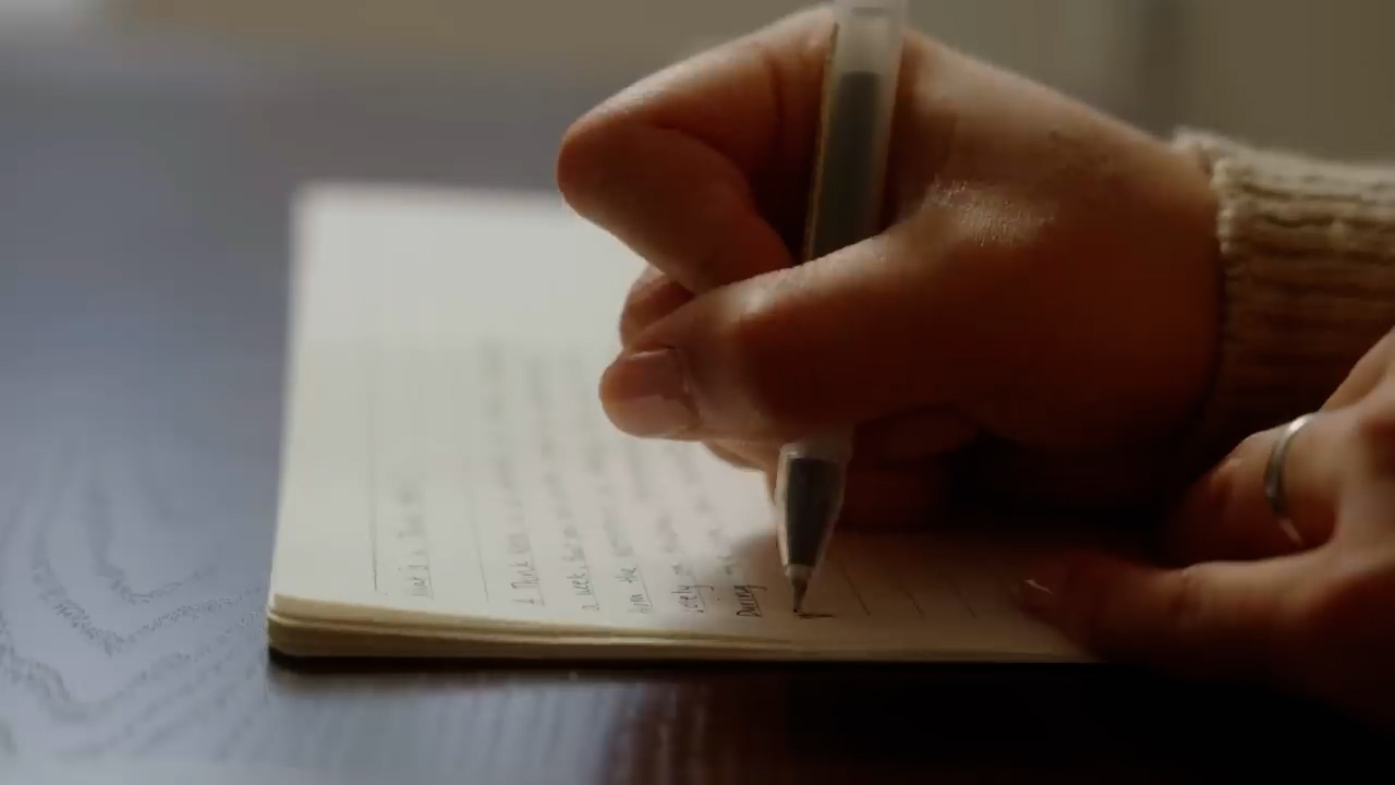 a closeup shot of a woman's hand writing in a notebook with a white pen. the notebook is open to a page with cursive writing visible. the scene is lit by soft, natural light coming from the left side, creating a warm and inviting atmosphere. the focus is on the hand and pen, emphasizing the act of writing.