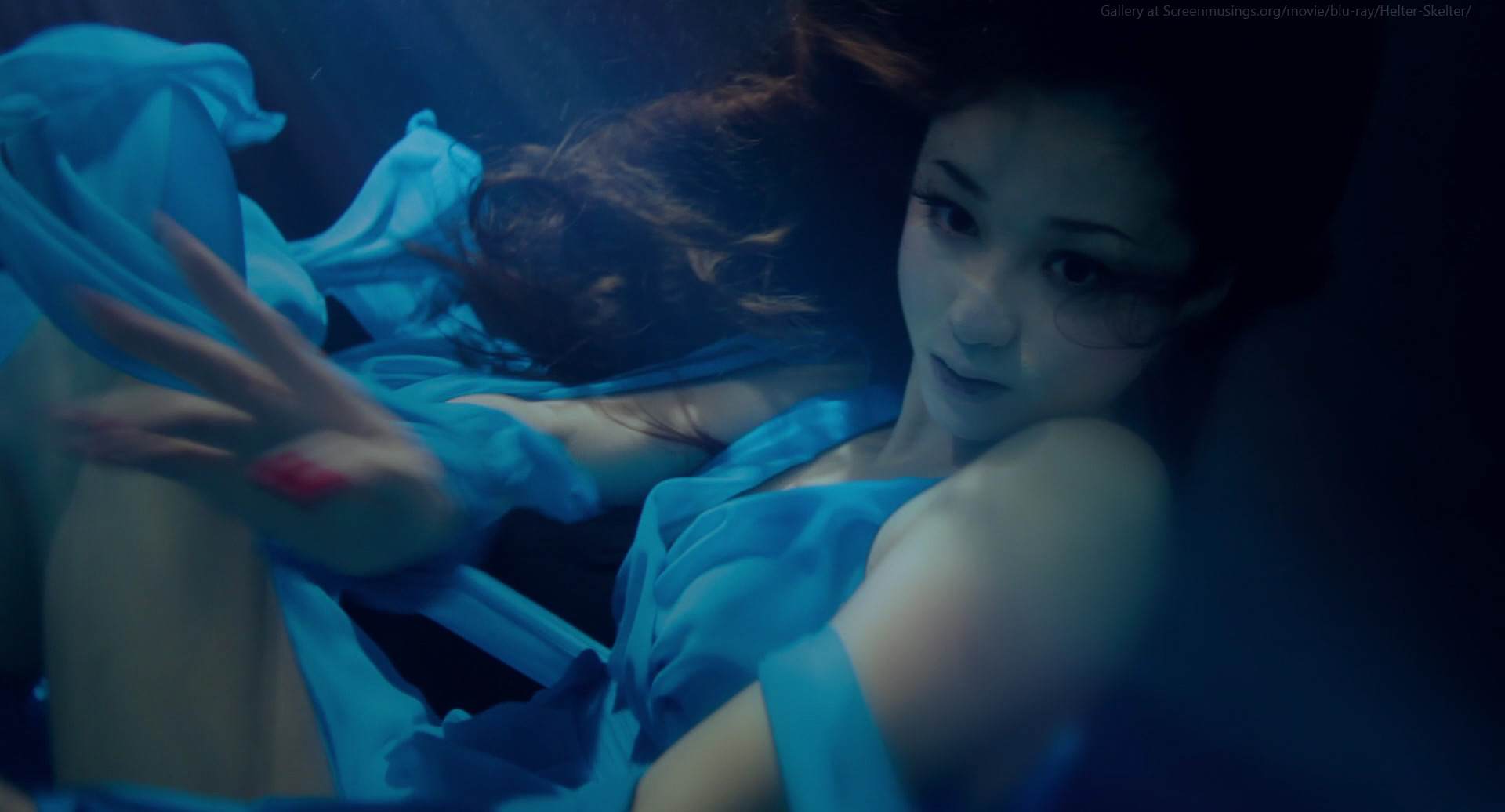 a woman with long dark hair and a blue dress is underwater in a studio shoot. her eyes are open and looking directly at the camera, giving an ethereal and mysterious feel. the lighting and editing creates a fantasy like feel.