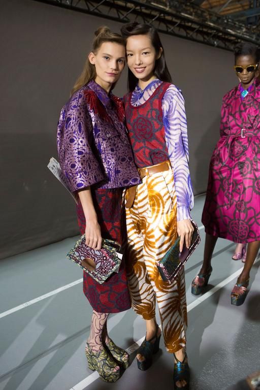 two models are standing close together, showcasing their outfits in a backstage setting. both models are wearing vibrant patterned clothing. the model on the left is wearing a purple brocade jacket and a dark red patterned skirt, while the model on the right sports a light purple and white patterned top with gold and white patterned pants.  both are wearing platform shoes with patterned fabric. behind them, another model in a pink floral dress is visible. the overall setting appears to be a fashion show or photoshoot. the models' expressions and poses convey confidence and poise. the focus is on the detail of the clothing and the overall fashion statement.