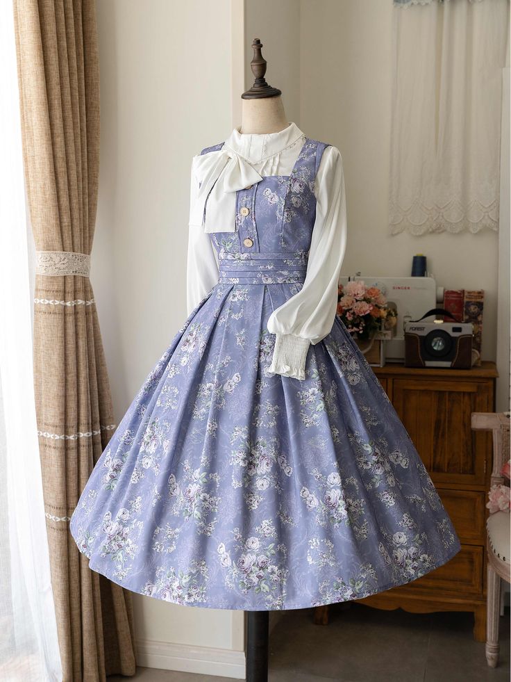 a fulllength photo of a light purple floral print lolita fashion dress on a mannequin. the dress features a square neckline, buttonup front, and a full, pleated skirt. it is paired with a white blouse with a large bow. the background is a white room with a window and a wooden cabinet.