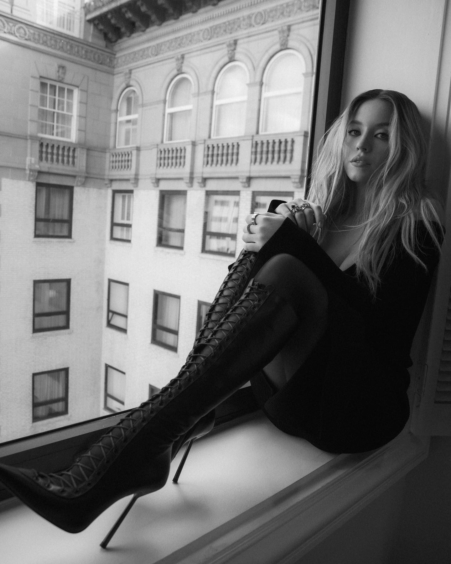 a black and white, fullshot photograph features a young woman with long blonde hair sitting on a window seat. she's wearing a black dress and very high, black laceup boots with stiletto heels.  her legs are crossed, and she's wearing rings. the window offers a view of a tall city building across the street. the overall mood is stylish and elegant. the image has a somewhat moody and dramatic feel due to the black and white contrast and the woman's pose.