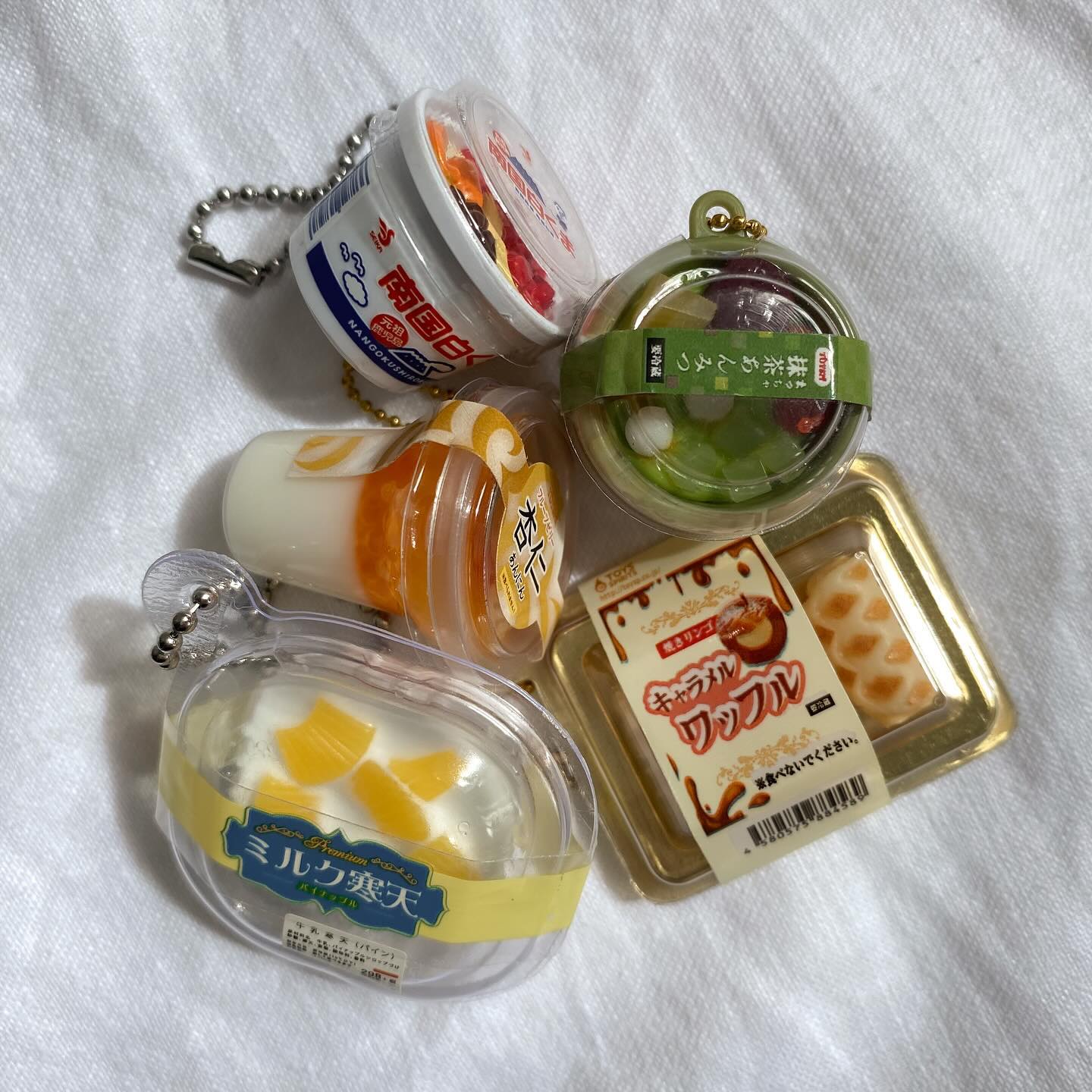 the image shows a collection of miniature dessert keychains, mostly japanese desserts. there is a white tub labeled "nankoku shirokuma," a clear container with mango pieces labeled "ミルク寒天" (milk jelly), a container with almond pudding labeled "杏仁" (almond), a round container of matcha anmitsu labeled "抹茶あんみつ" (matcha anmitsu), and a packaged caramel waffle labeled "キャラメルワッフル" (caramel waffle).