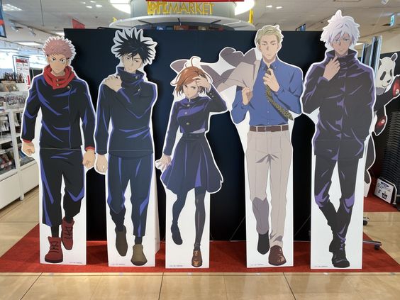 this image shows five lifesize standees of characters from the anime jujutsu kaisen, lined up against a back wall in what appears to be a retail store. the characters are: yuji itadori, megumi fushiguro, satoru gojo, nobara kugisaki, and kento nanami. the standees are positioned in front of a promotional wall, and the store's ceiling with fluorescent lighting is visible in the background.  each character is depicted in their signature outfits, and the overall aesthetic of the image is bright and colorful, capturing the essence of the popular anime series.