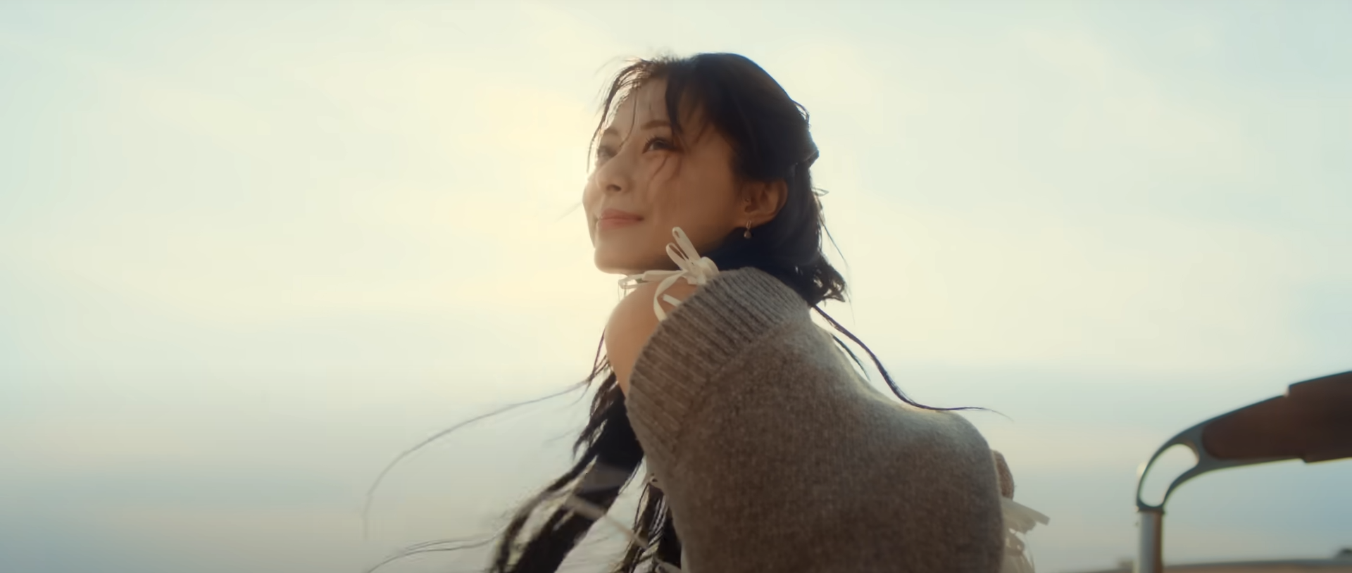 woman with long dark hair is shown in profile, her face turned towards the sun. she is wearing a loosefitting, knitted, offtheshoulder sweater in a light brown color. the sunlight creates a bright backlight, making her hair glow and outlining her figure. the background suggests a beach setting with a clear sky and hints of sand and a beach umbrella in the lower right corner. the overall impression is one of warmth, happiness, and carefree summer vibes.