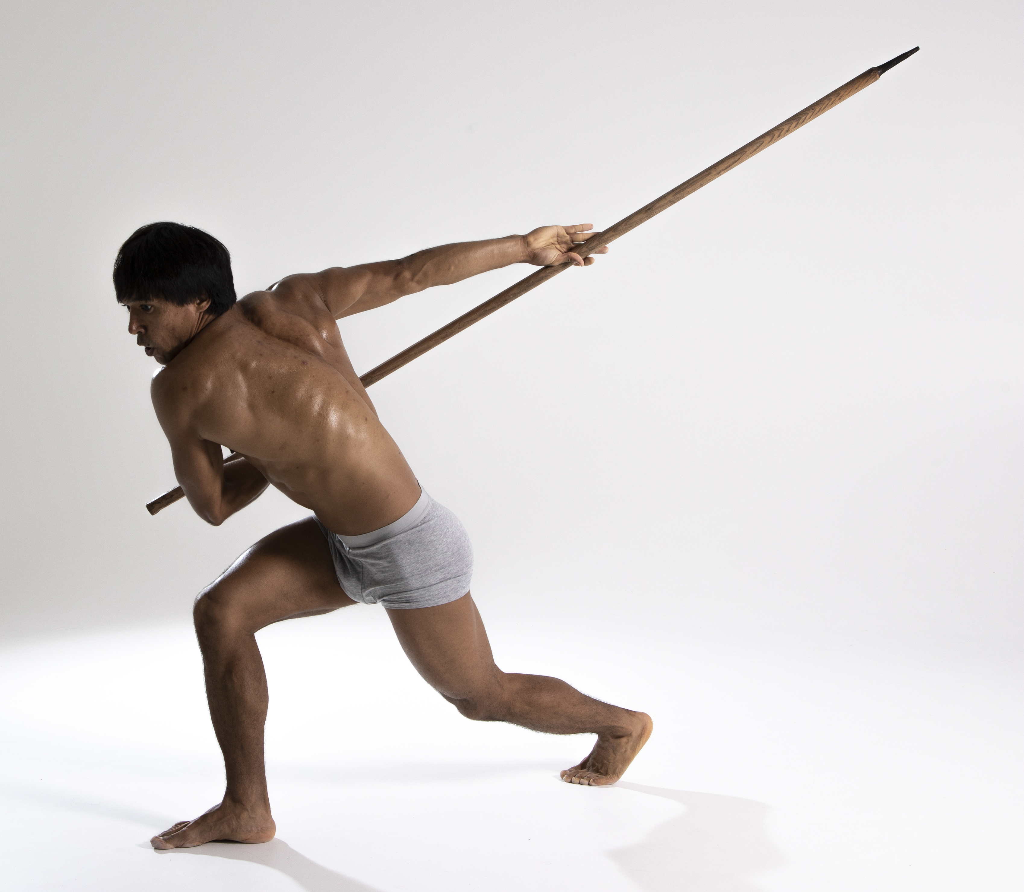 a muscular, shirtless man in a loincloth stands barefoot in a studio, poised to throw a spear. his body is oiled, and he is positioned in a dynamic stance with one leg forward and the other extended behind him, ready to launch the spear with force.