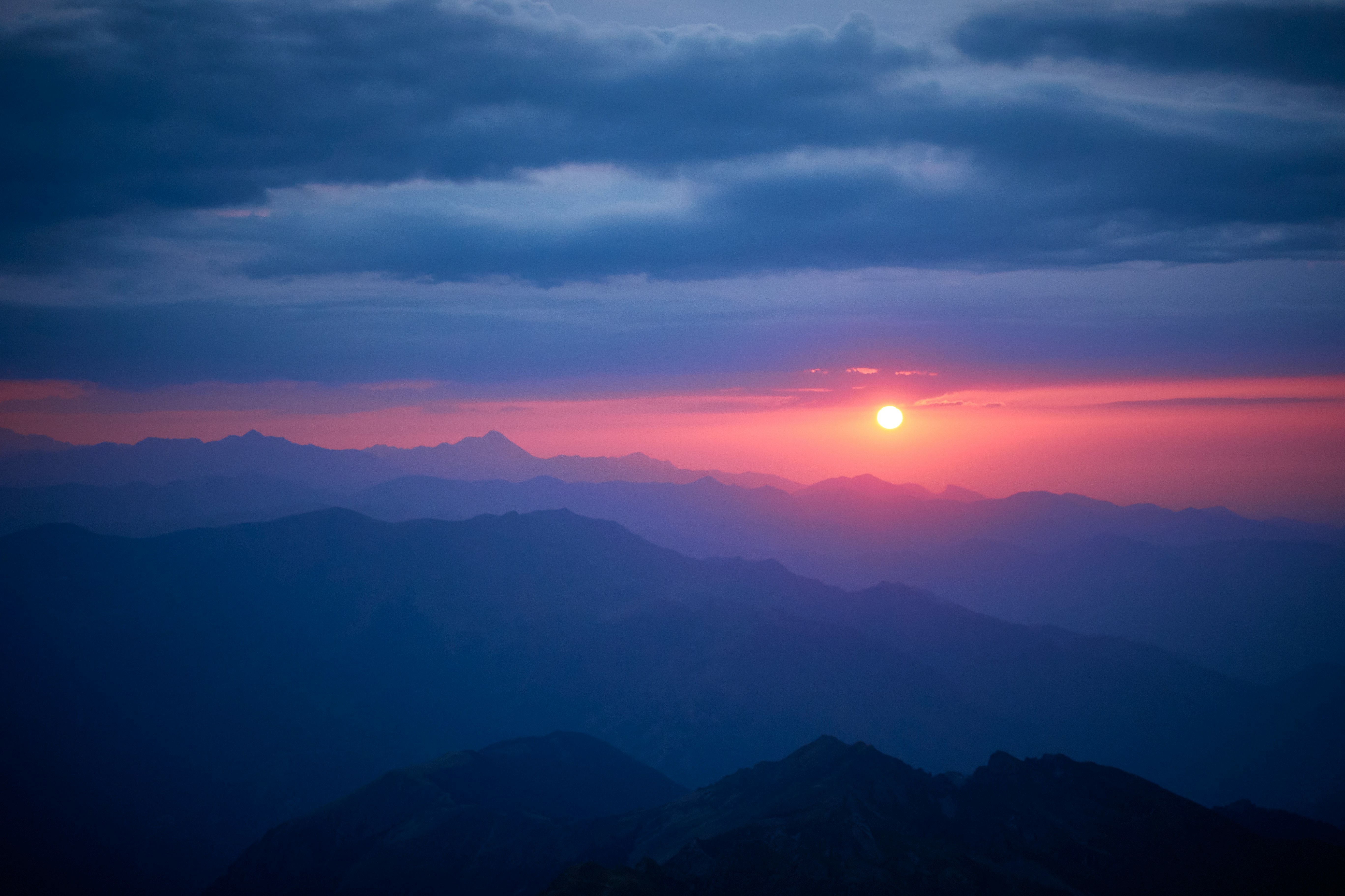 the image captures the breathtaking beauty of a mountain range at sunset. the sun, a fiery orb, dips below the horizon, casting an ethereal glow across the sky in hues of orange, pink, and purple. the mountains, silhouetted against the vibrant sky, create a dramatic and majestic scene. the clouds, painted in shades of blue and gray, add depth and texture to the composition. the overall effect is one of tranquility and aweinspiring beauty.