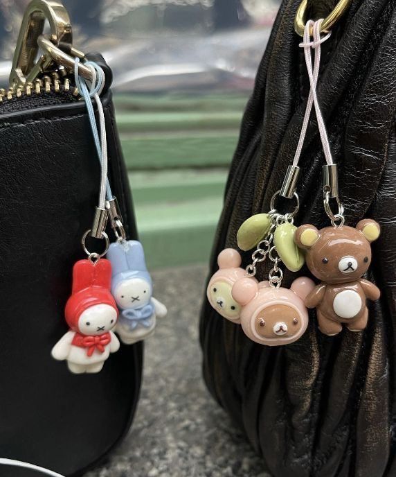 the image shows two black purses, each adorned with cute character keychains. the purse on the left has a my melody and a bunny charm. the purse on the right has rilakkuma bear charms and small green leafshaped charms.