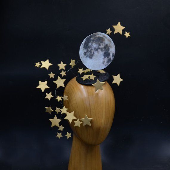 a mannequin head is adorned with a black hat featuring a large paper moon and numerous gold star decorations against a black background.