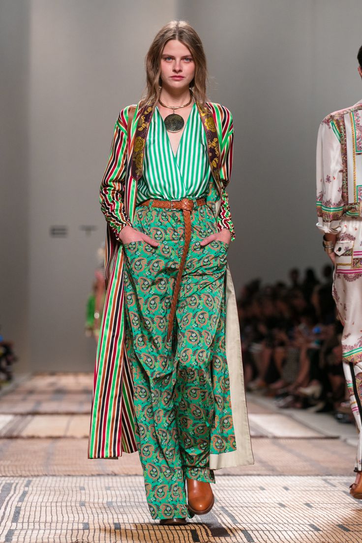 a fashion model walks down a runway, showcasing a vibrant green paisley print pantsuit.  the outfit includes a green and white striped top under a long, multicolored striped coat.  she is accessorized with a large, circular necklace and brown ankle boots.  another model in the background is partially visible, wearing a different patterned outfit. the runway is a neutral tone, allowing the model's outfit to stand out.  the overall style suggests a bohemian or 70sinspired look.