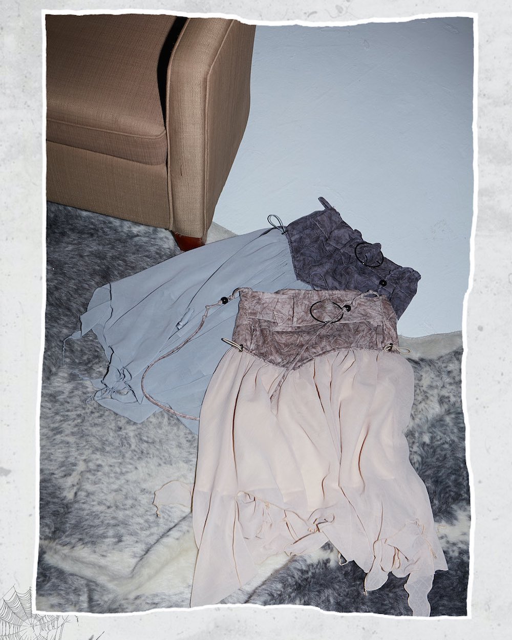a closeup shot showcasing three skirts layered on top of each other.  the top skirt is a dark gray, possibly suede or a similar textured fabric.  underneath is a light blue sheer skirt, followed by a light beige, flowy skirt at the bottom. the skirts are loosely draped and appear to have a vintage or bohemian style. they are arranged on a gray and white patterned rug near a beige armchair, creating a soft, elegant stilllife composition. the overall color palette is muted and neutral.