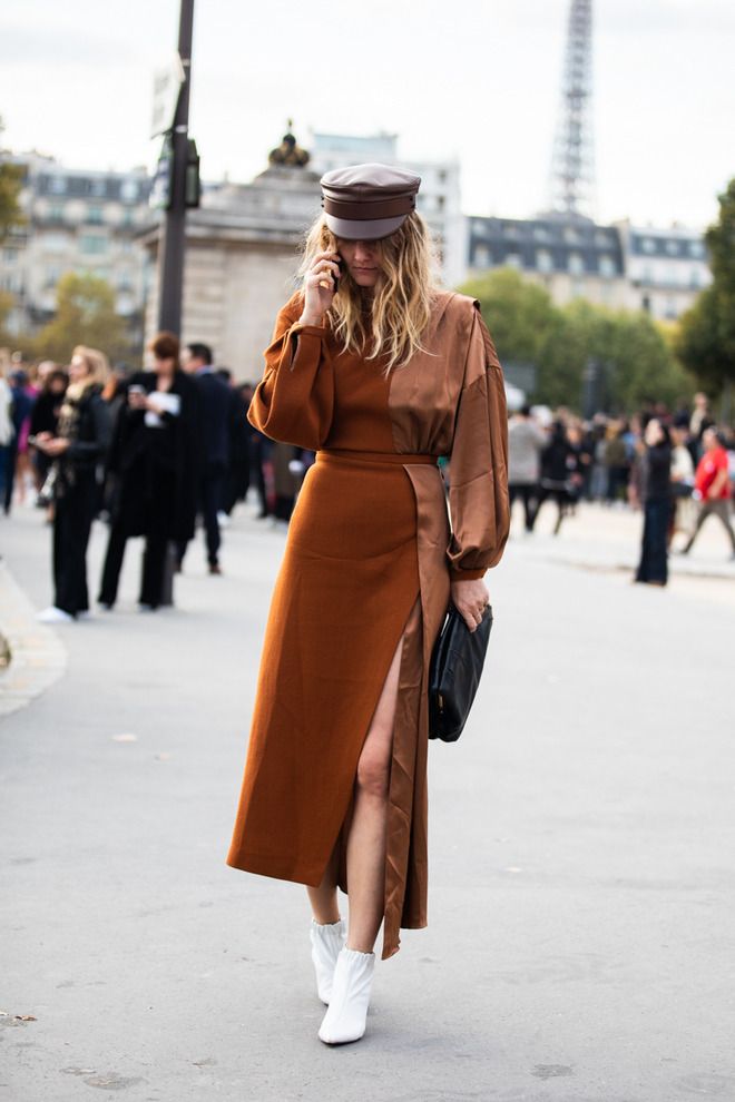 the image shows a stylish woman in a rustcolored midi dress with a side slit and a brown silk overlay detail.  she's accessorized with a brown leather newsboy cap, white ankle boots, and a black clutch. she is on a parisian street, with the eiffel tower subtly visible in the background. the woman is on a phone call. the overall style is autumnal and chic, reflecting current street style trends. the photo is likely a candid street style shot during fashion week or a similar event.