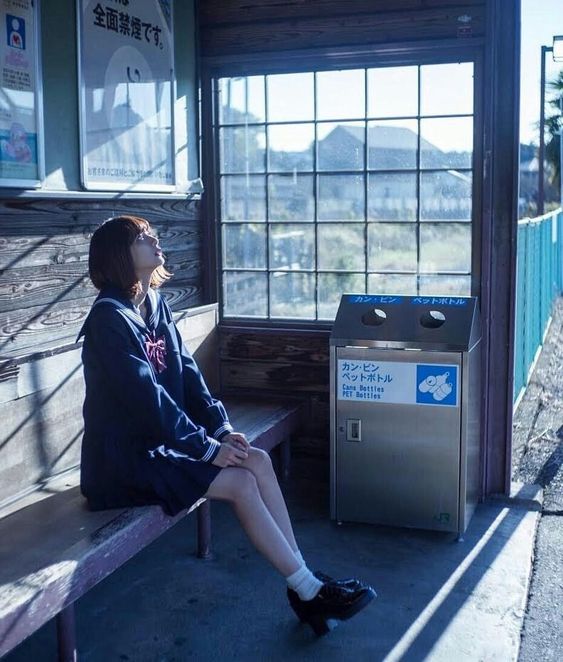 a young woman in a japanese school uniform sits on a bench at a rural train station, bathed in soft sunlight. she gazes off to the side, lost in thought as she waits. the image evokes a sense of quiet contemplation and the passage of time. the color palette is muted and cool, emphasizing the serene atmosphere.