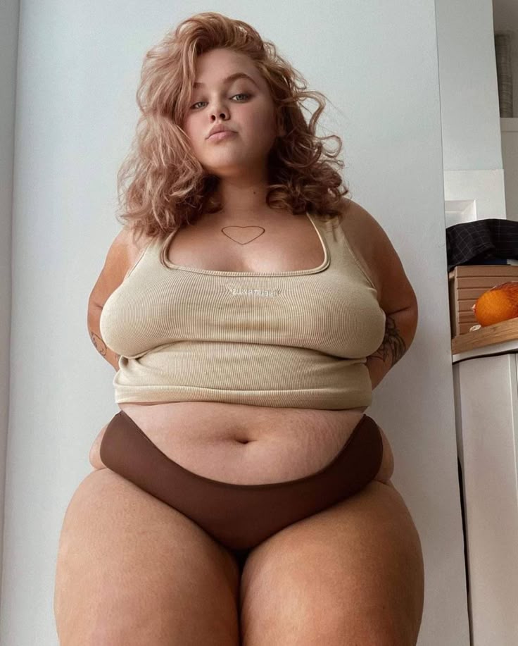 a woman with blonde curly hair is wearing a tank top and underwear. she has a heart tattoo on her chest. she has thick thighs and a curvy figure.