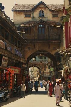 the image showcases a bustling street scene in india, likely a market area, teeming with people going about their day. the perspective is from street level, drawing the viewer directly into the heart of the action. traditional indian architecture lines the street, characterized by ornate balconies and archways. sunlight bathes the scene, casting long shadows and creating a vibrant atmosphere. the warm color palette reinforces the sense of energy and life.