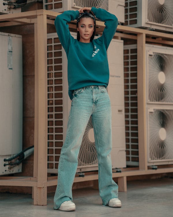 the image shows a young woman standing outdoors against a background of stacked air conditioning units. she's wearing a tealcolored crewneck sweatshirt with subtle branding, light blue wideleg jeans, and white sneakers.  her hair is pulled back with a headband.  she's posing casually, hands behind her head, showcasing a relaxed yet stylish outfit. the setting appears to be an urban area, with the air conditioning units indicating a building exterior or possibly a warehouse backdrop. the overall style is casual and comfortable, indicative of current fashion trends.