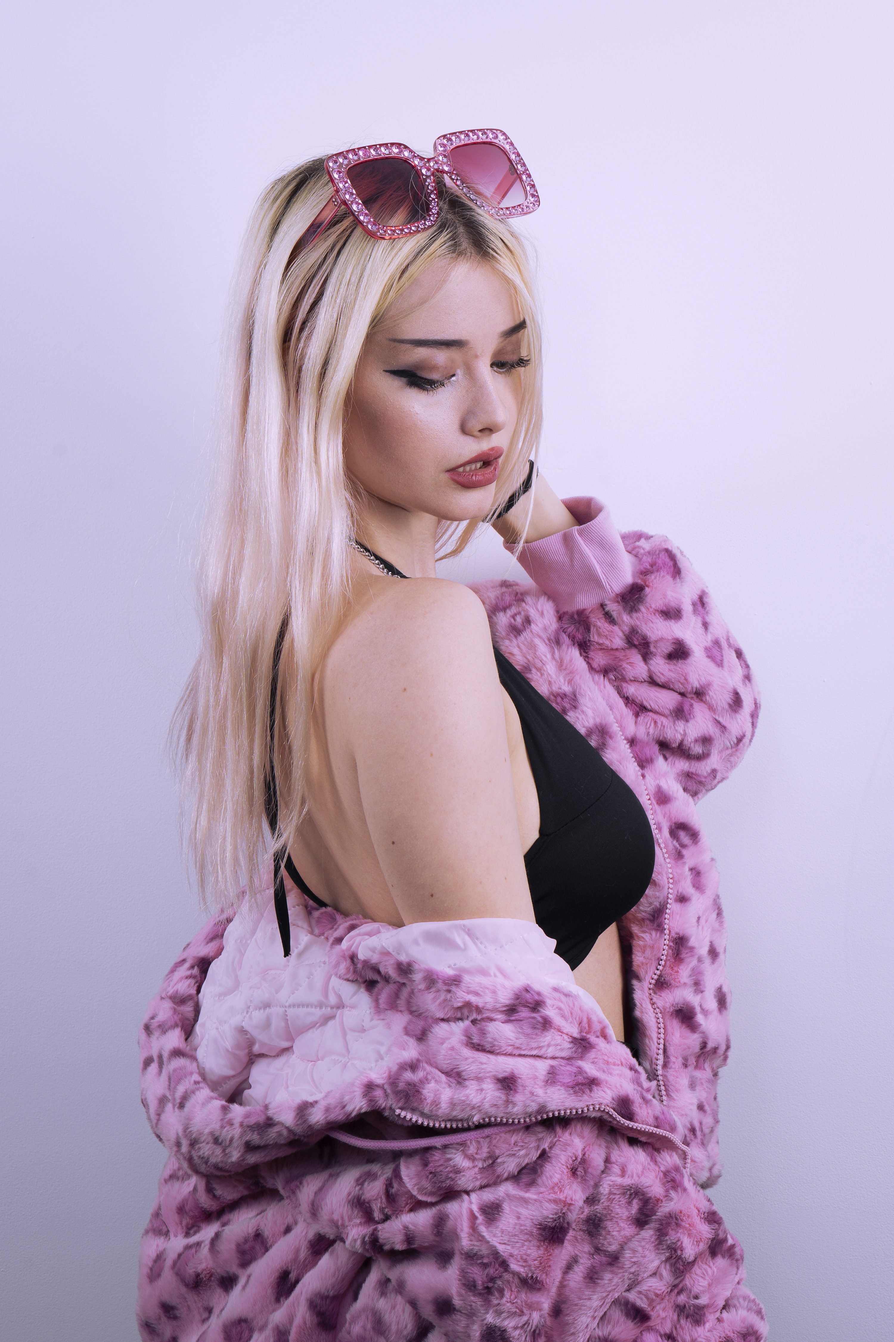 a closeup shot of a young blonde woman posing against a light pink background. she's wearing a fluffy, pink leopard print jacket, a black bralette, and pink jeweled sunglasses perched on her head. her hair is shoulder length and styled softly. she looks downwards, giving a slightly melancholic or pensive expression. the image focuses on her clothing and style, creating a fashionforward and slightly edgy aesthetic. the soft lighting and the color palette contribute to a dreamy and stylish feel.