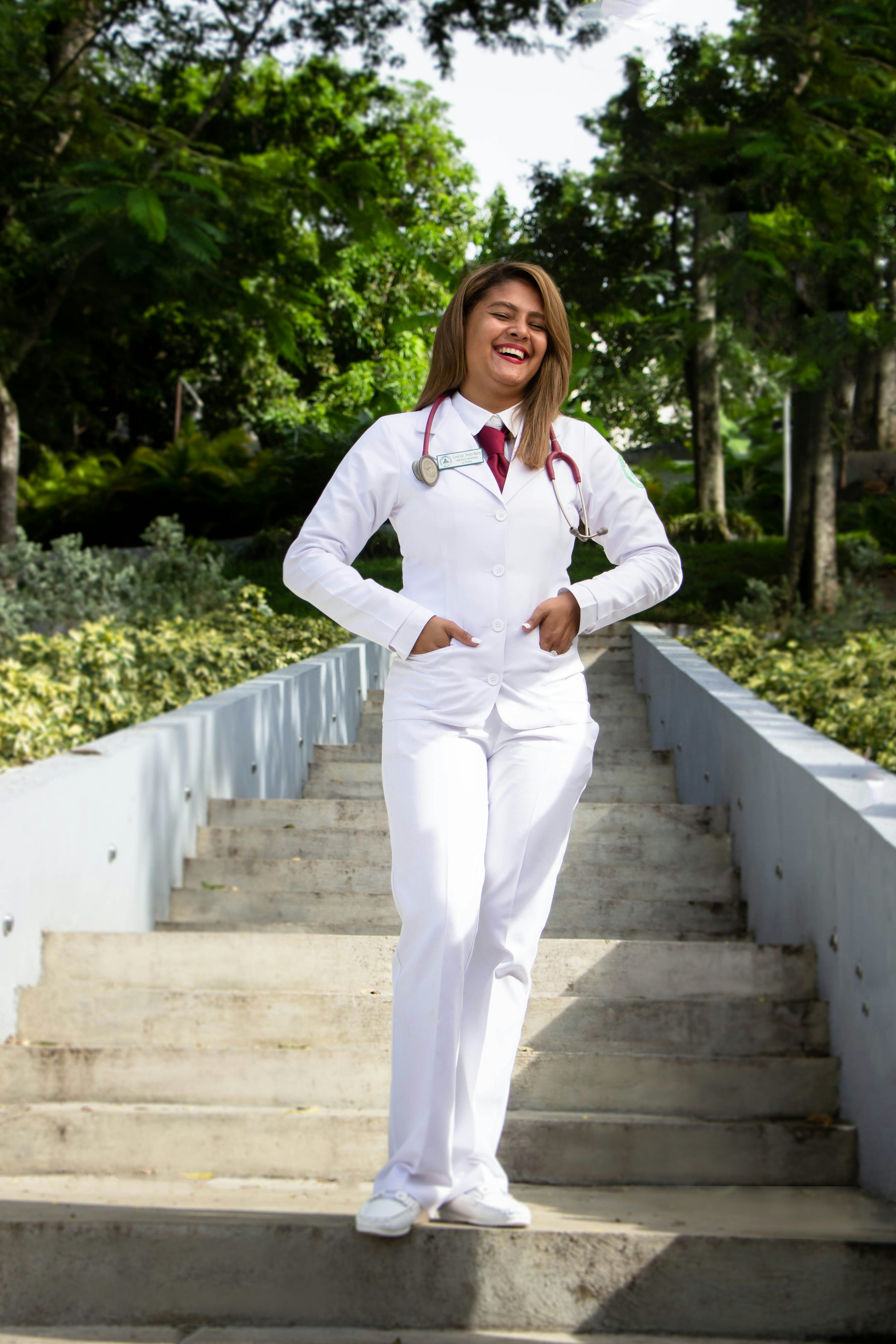 a young latina woman, dressed in a white doctor's coat and stethoscope, stands happily on a set of outdoor stairs.  she is smiling and has her hands in her pockets.  the background is a lush green park with trees and sunlight. the overall image projects a feeling of accomplishment and hopefulness.