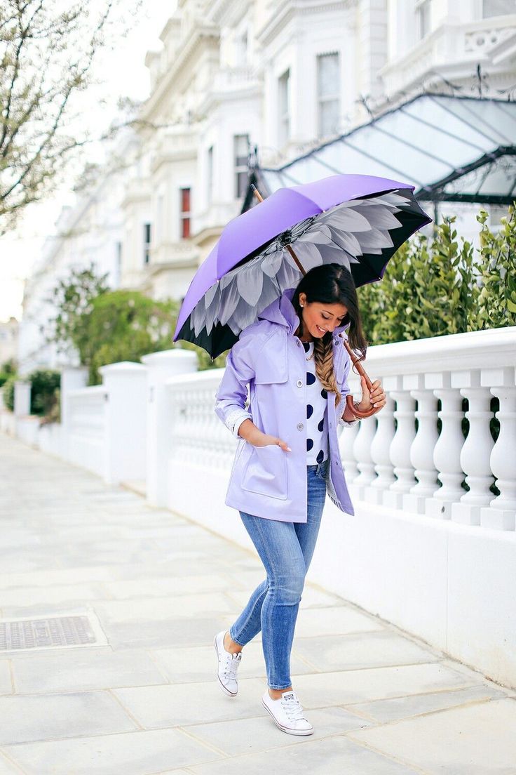 a stylish woman is walking down a city street, carrying a purple umbrella, wearing a purple jacket, and white shoes. she is looking down at the ground as she walks. there is a white fence on the right side of the image. the sky is overcast.