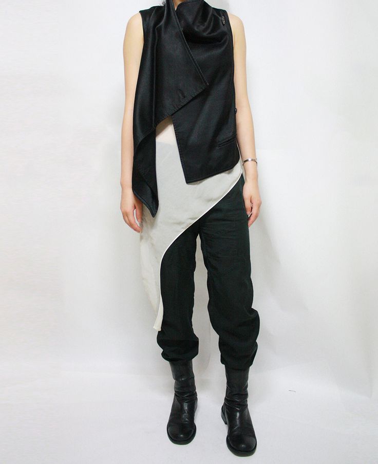 a person is standing in front of a white background wearing a sleeveless black vest with a large draping collar, a sheer offwhite blouse, dark green cuffed pants, and black leather boots.