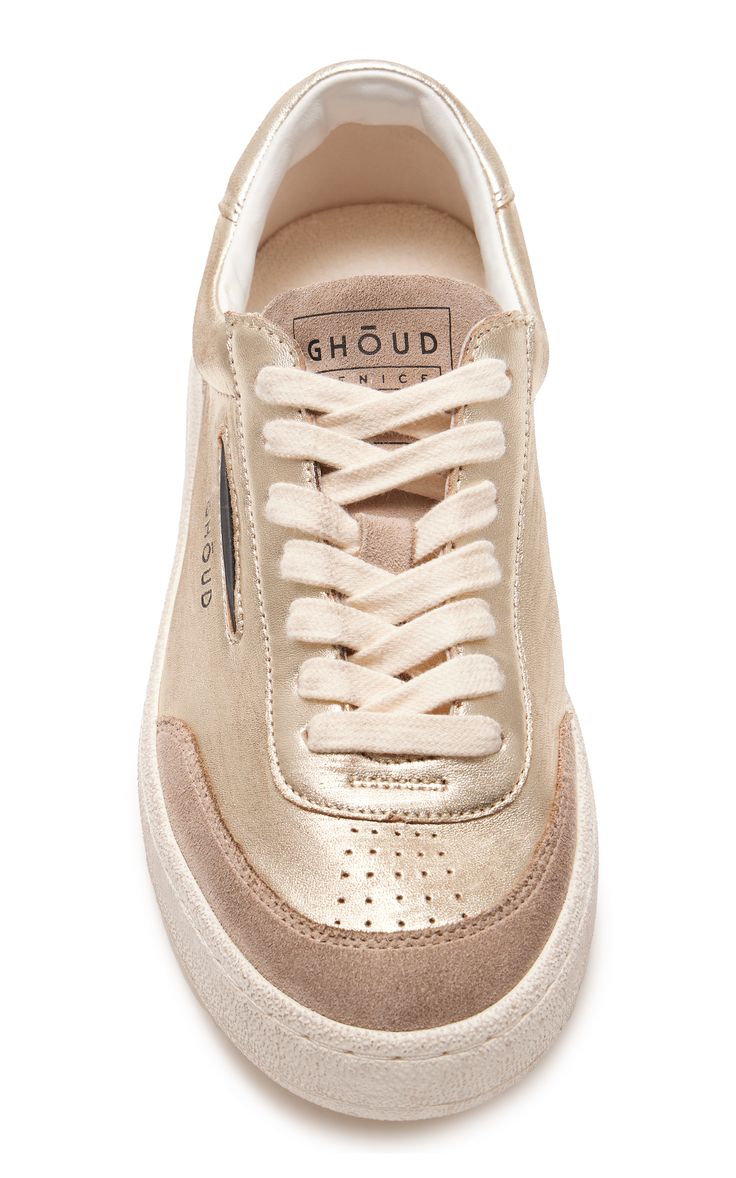 a closeup studio shot of a pair of women's ghoud sneakers in a metallic gold color. the shoes are lowtop sneakers with beige laces and a creamcolored sole.  the ghoud branding is visible on the tongue.  the image shows the shoes against a white background.