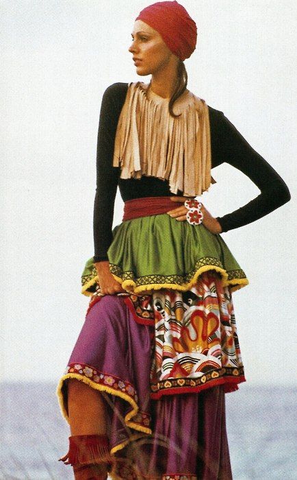 a woman in 1970s fashion is posing for the camera. she is wearing a red turban, black longsleeved top with a fringe collar, a green skirt, and a purple overskirt. the overskirt is decorated with a colorful pattern. the model is looking to the right of the frame.