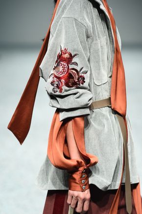 loseup shot of a woman's upper body showcasing a detailed grey shirt with orange embroidery on the sleeves. the shirt has a relaxed fit and features a unique design with a combination of fabrics and textures. an orange scarf is loosely draped around the neck and shoulders, adding a pop of color to the overall look. the model is wearing the shirt as part of a runway or fashion show presentation, highlighting its design and style. the focus is on the intricate embroidery and the interplay of colors and textures in the garment.