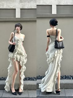 a woman in a white ruffled dress with a high slit and heels. she is posing outdoors and carrying a black bag. the woman is in a chic and stylish outfit.