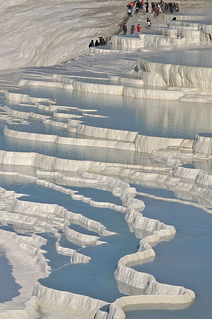 the image showcases the stunning white calcium terraces of pamukkale, turkey, filled with blueturquoise hot spring water. several people are visible walking along the top terraces, enjoying the unique natural landscape. the stepped formations create a visually striking scene of white against the blue water.
