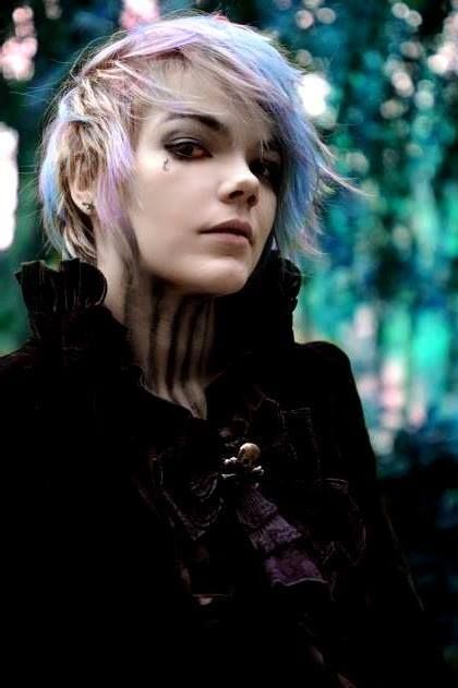 a portrait of a woman with short, multicolored hair, wearing a black gothicstyle jacket with a high collar. she has pale skin, dark eye makeup, and a teardropshaped tattoo below her left eye. her neck has dark, vertical markings resembling stripes. the jacket features a skull detail. the background is blurred, suggesting an outdoor setting with trees and foliage.
