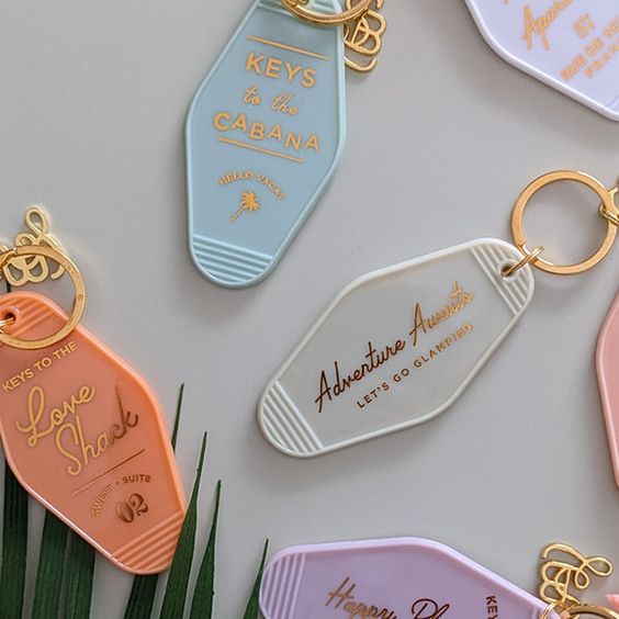 a flat lay of five different colorful keychains. each keychain is shaped like a motel key and features a different goldlettered message. they are displayed against a light gray background, with a few green palm leaves peeking in from the bottom of the frame.