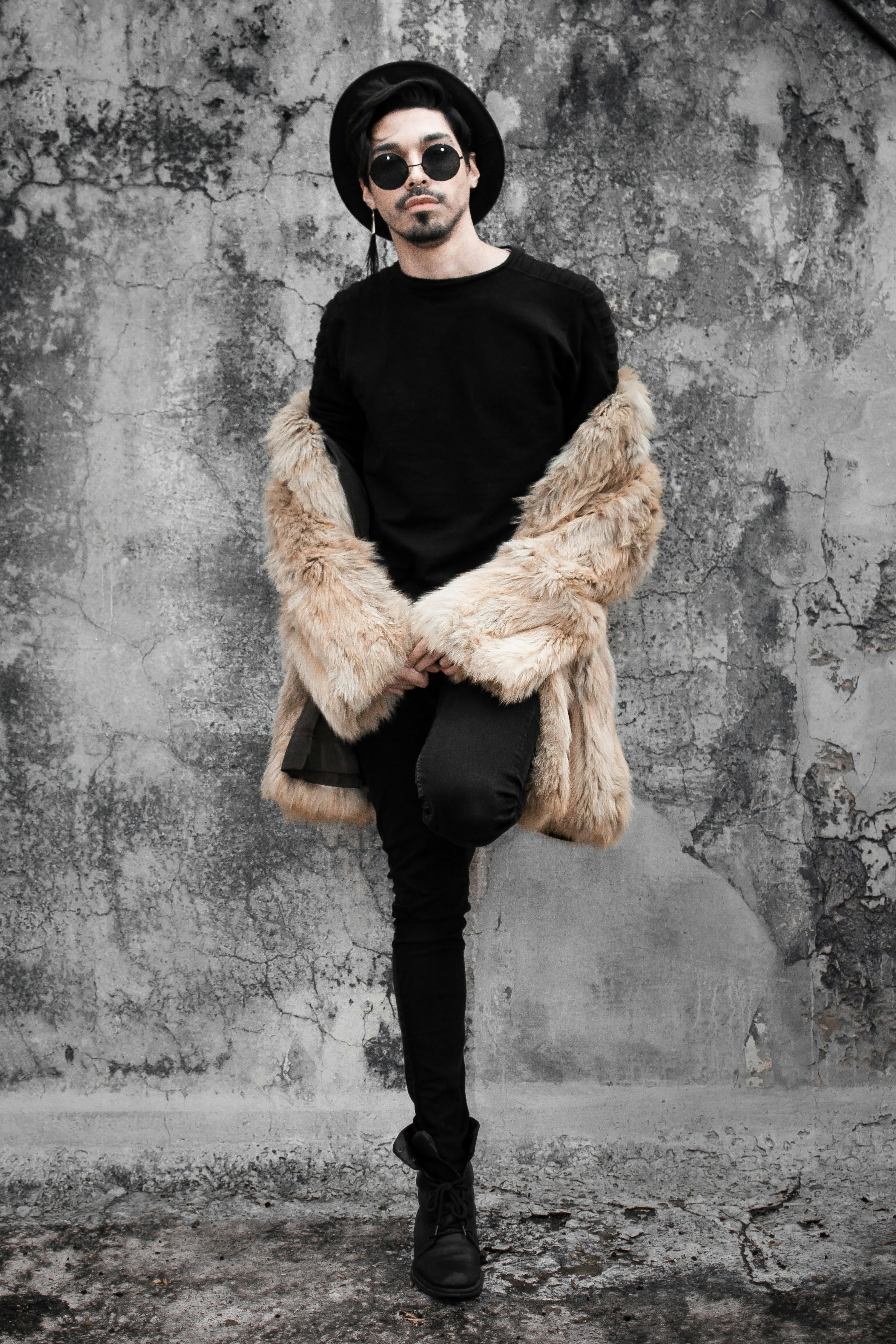 a full shot of a stylish man posing against a grunge concrete wall. he is wearing a beige faux fur coat over a black shirt, black skinny jeans, black boots, sunglasses and a black fedora hat. his pose is relaxed and confident, showcasing his fashion sense. the image has a cool, trendy and slightly hipster vibe. the monochrome color scheme enhances the overall mood.