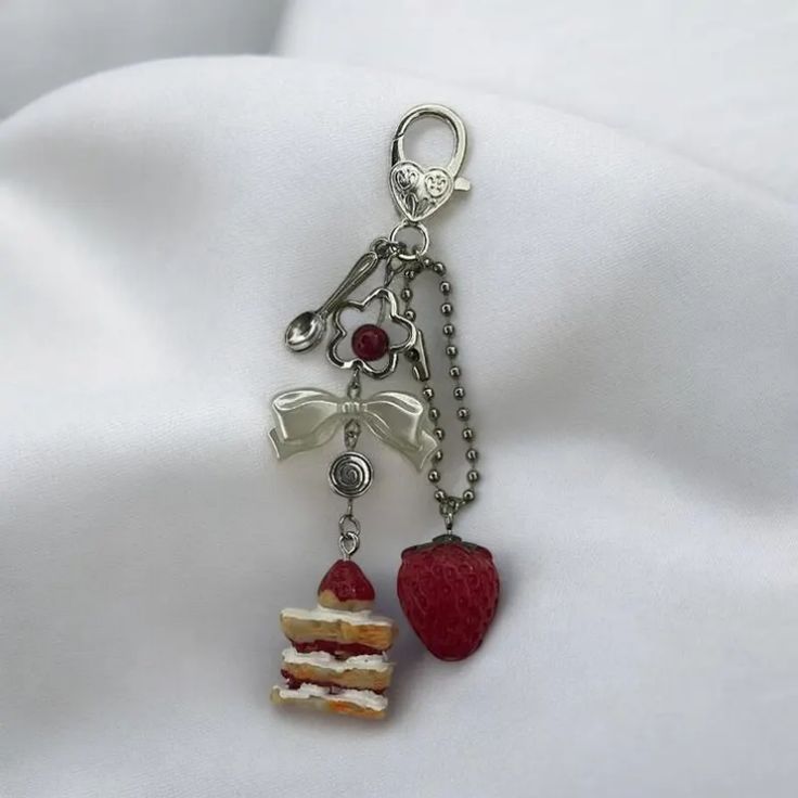 a charm keychain hangs over a white fabric background. the keychain consists of a silver clasp with a heart design at the top, followed by a silver spoon charm, a flower charm with a red bead at the center, a translucent white bow charm, a silver swirl charm, a miniature cake charm, a strawberry charm hanging from a ball chain, and a small silver ring connecting the cake and swirl charms. the keychain's charms have a dessert and trinket theme.