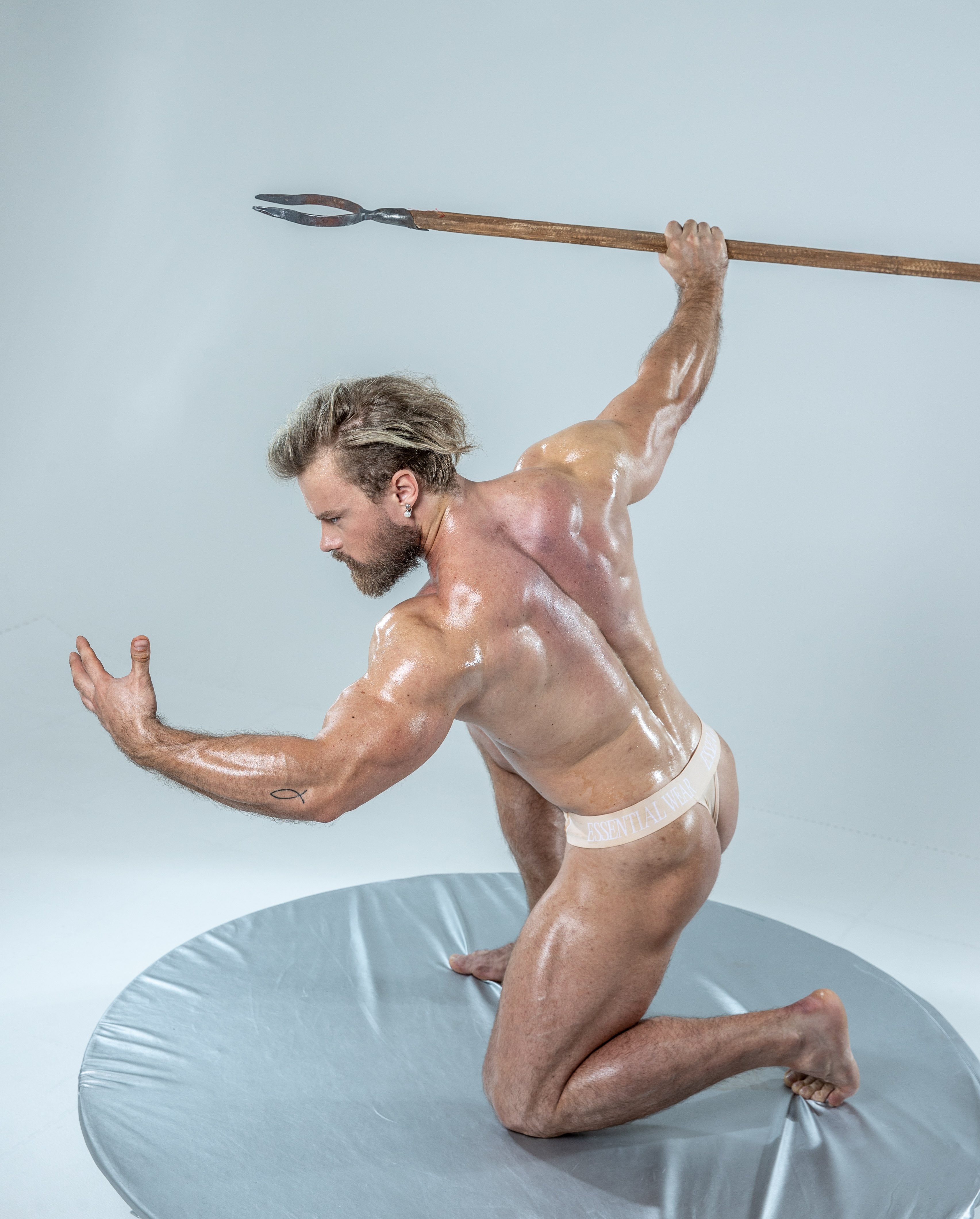 a muscular man with oiled skin stands on a gray platform, holding a spear. he is positioned with his back to the camera, his body slightly twisted, and his head turned to the left. his muscles are tense, highlighting his physique. he wears only a white underwear. the backdrop is a simple gray, suggesting a studio setting.