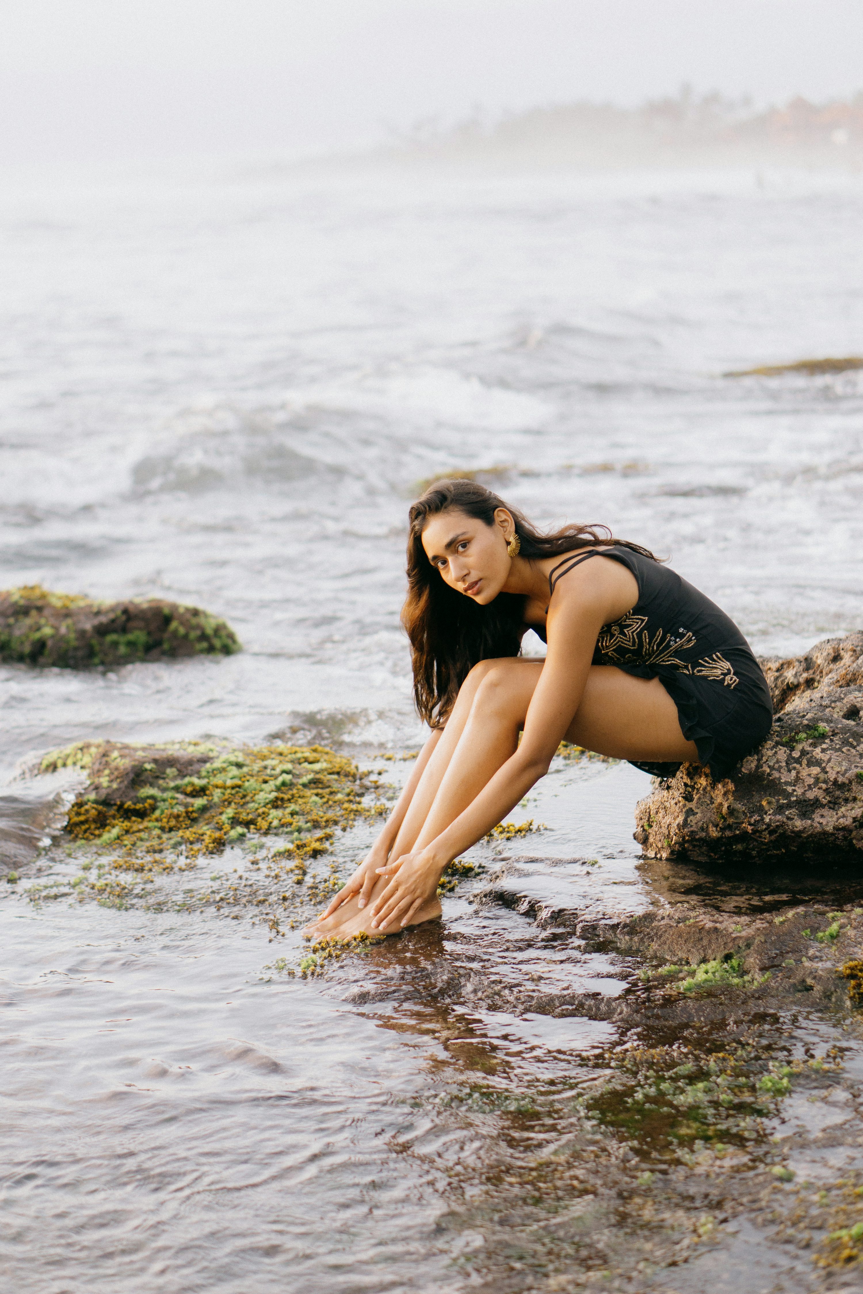 a young woman with long dark hair sits on rocks at the edge of the ocean, her legs dangling in the shallow water. she is wearing a darkcolored dress and gold earrings. the scene is peaceful and serene, suggesting a relaxing vacation or holiday. the ocean is calm, and the rocks are covered with some green moss.  the overall mood is one of tranquility and beauty.
