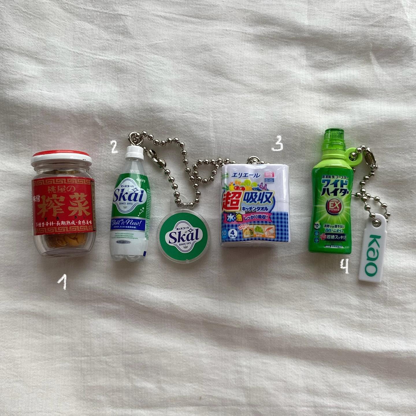 the image shows four keychains lined up on a white surface. the first keychain is a miniature jar of momoya preserved mustard greens. the second keychain consists of a small bottle of skal soda and a round skal logo pendant. the third keychain is a packet of erika super absorbent kitchen towels. the fourth keychain is a bottle of kao wide haiter ex stain remover and a kao tag.