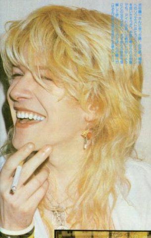 a closeup portrait of a woman with big 80s blonde hair smiling brightly and holding a lit cigarette. she's wearing multiple necklaces and earrings.  the image has a vintage, grainy quality.