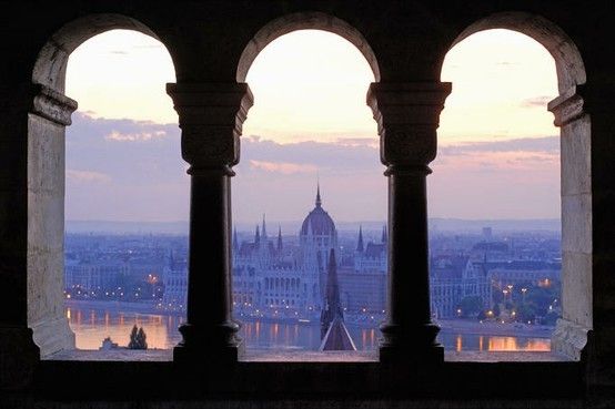 the image is a captivating view of the budapest cityscape, specifically featuring the hungarian parliament building, seen through three arched windows. the scene is bathed in the soft, pastel hues of either sunrise or sunset, creating a tranquil and romantic atmosphere. the architectural elements of the framing structure – the stone arches and columns – add to the image's elegance. the danube river is visible in the foreground, reflecting the city lights and the soft sky. the overall composition offers a balanced blend of urban landscape and architectural framing, capturing the essence of budapest at a magical hour.