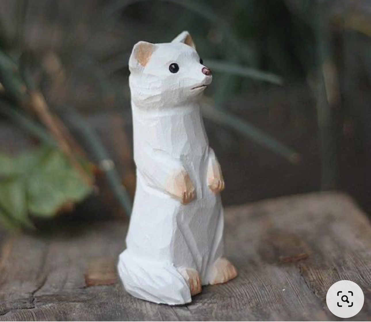 a white wooden figurine of a weasel or stoat stands on a surface that appears to be weathered wood.  the figurine is smooth and carved in a simple style, with minimal detail in the fur and face, but the pose and proportions of the animal are well captured. the figurine is mostly white with light brown highlights on the tips of the paws and ears, and black beads for the eyes. the background is blurred, indicating an outdoor setting, and the lighting is natural and soft.