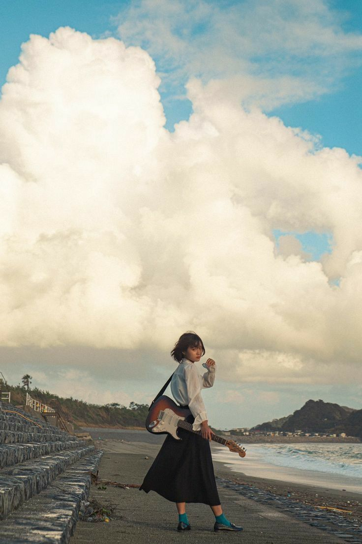 a young woman stands on a beach holding an electric guitar. she is wearing a white blouse, a long black skirt, and blue socks with shoes. she is looking back over her shoulder. the background consists of a cloudy sky, the ocean, and a small town on a hill in the distance.
