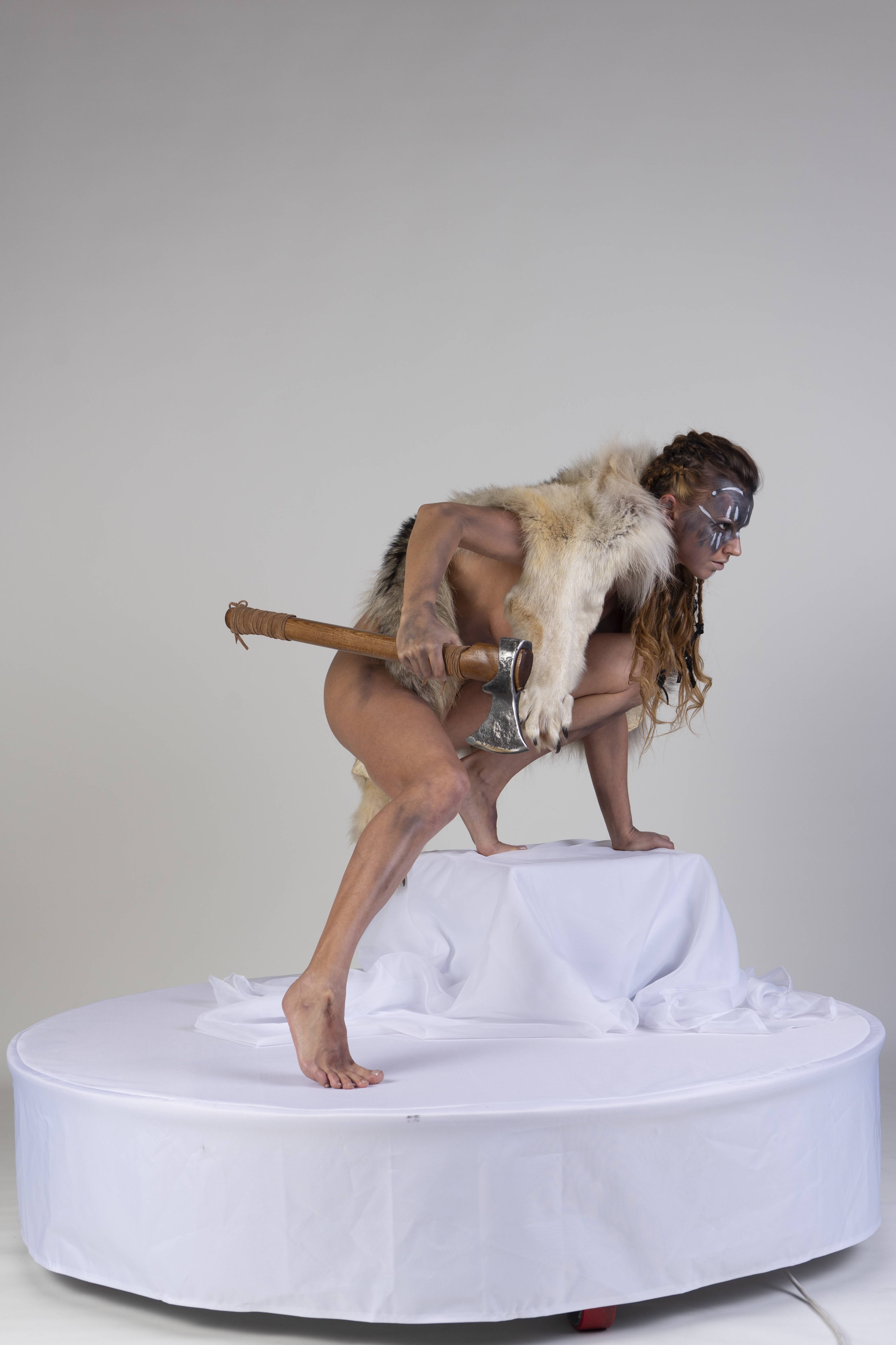 a woman dressed as a viking is squatting on a white platform, holding an axe. she is wearing a fur pelt and has braids in her hair. her face is painted with white markings. the background is a plain white studio backdrop.