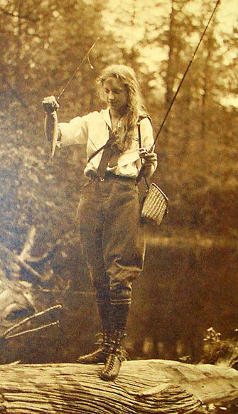 a woman in vintage clothing holds a fishing rod and a fish while standing on a log by a river. the image is in sepia tones.